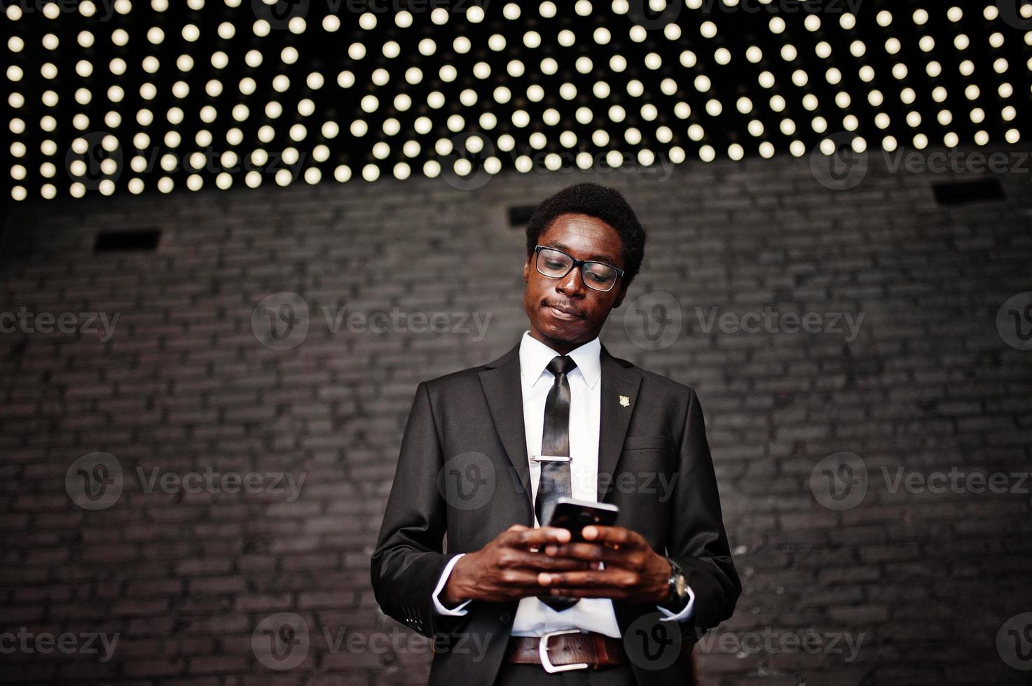 uomo d'affari afroamericano indossare su abito nero e occhiali in ufficio guardando sul telefono cellulare contro luci futuristiche. foto