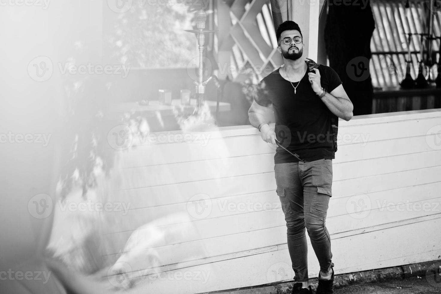 elegante barba uomo arabo in occhiali e t-shirt nera che fuma narghilè all'aperto. modello arabo che riposa. foto