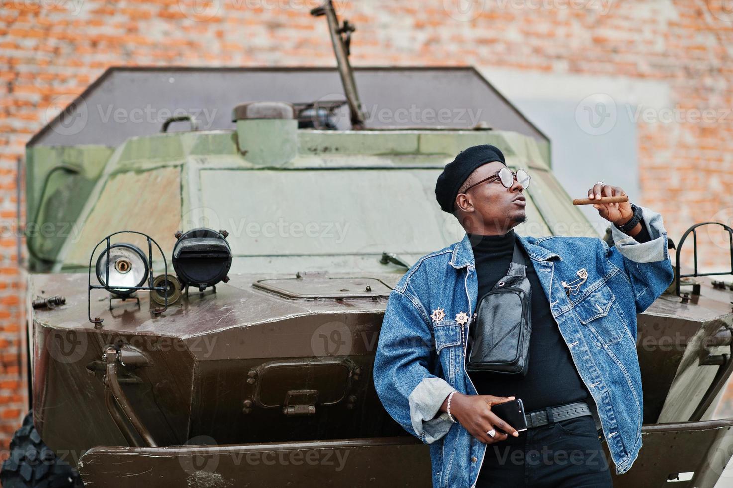 uomo afroamericano in giacca di jeans, berretto e occhiali da vista, con sigaro posato contro un veicolo blindato militare btr. foto