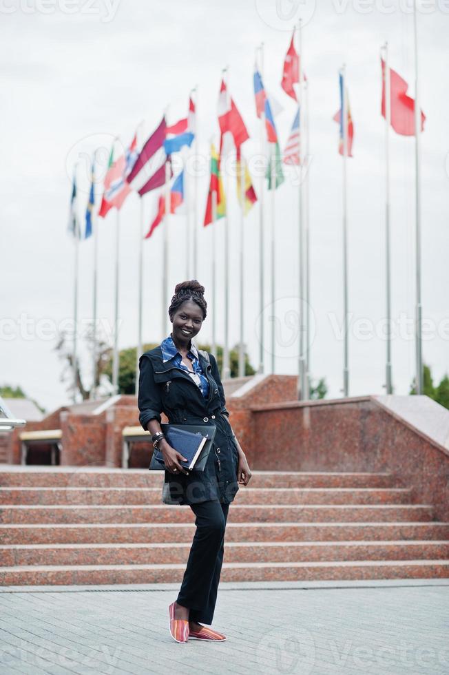 studentessa africana in posa con zaino e articoli per la scuola nel cortile dell'università, contro le bandiere di diversi paesi. foto