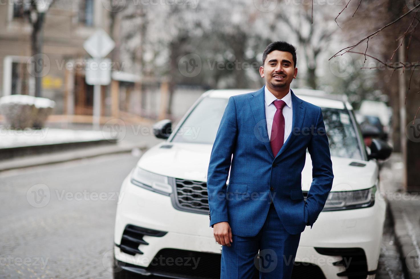 elegante modello indiano uomo alla moda su abito posato al giorno d'inverno contro un'auto bianca business suv. foto