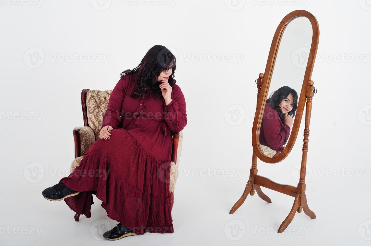 attraente donna del sud asiatico in abito rosso intenso poste in studio su sfondo bianco contro lo specchio e seduto alla sedia. foto