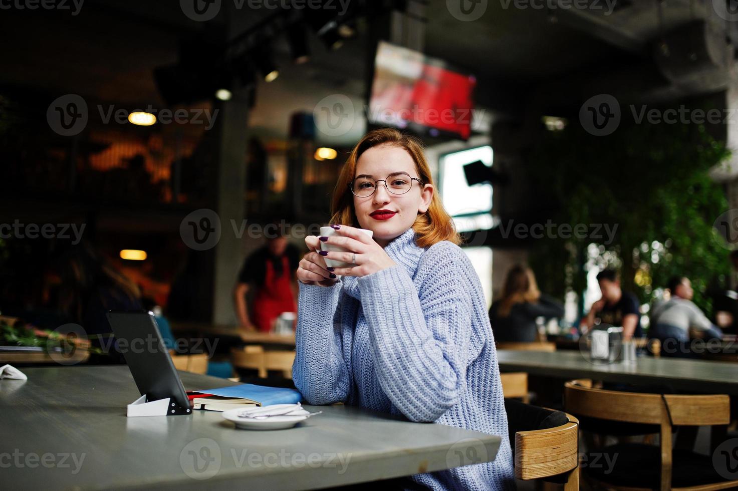 allegra giovane bella donna dai capelli rossi con gli occhiali usando il suo telefono, touchpad e notebook mentre era seduto al suo posto di lavoro al bar con una tazza di caffè. foto