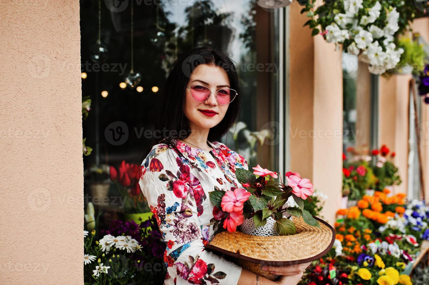 ritratto estivo di ragazza bruna con occhiali rosa e cappello contro il negozio di fiori. foto