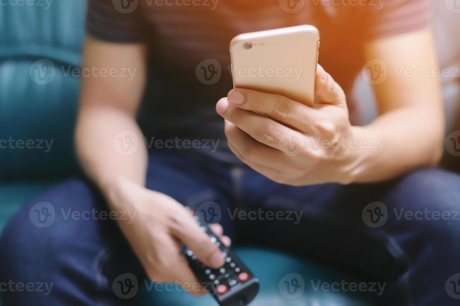 il giovane che utilizza il telecomando della televisione e lo smartphone mobile è seduto su un divano. acceso o spento tv. foto