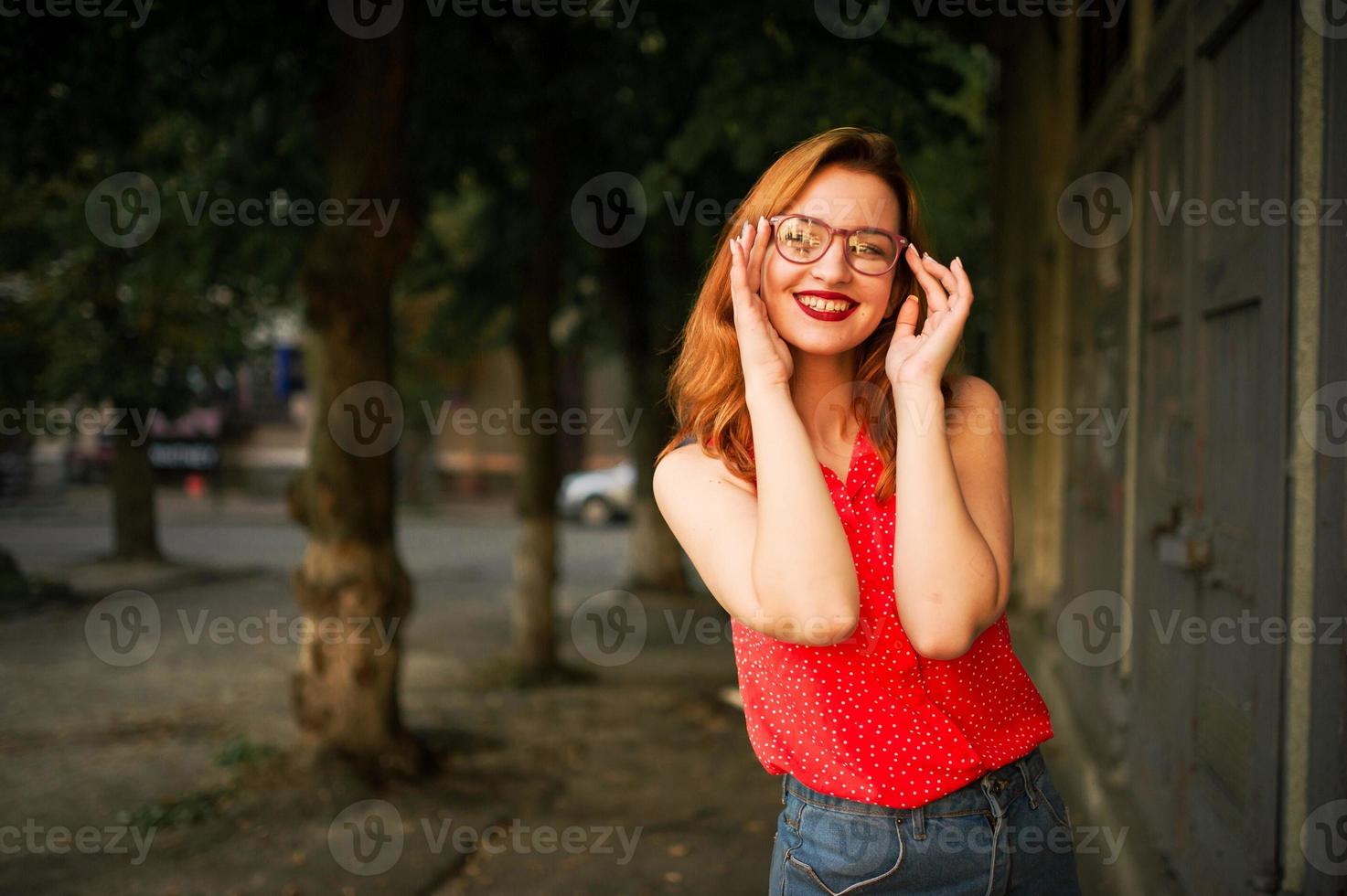 attraente donna dai capelli rossi in occhiali, indossare su camicetta rossa e gonna di jeans in posa. foto