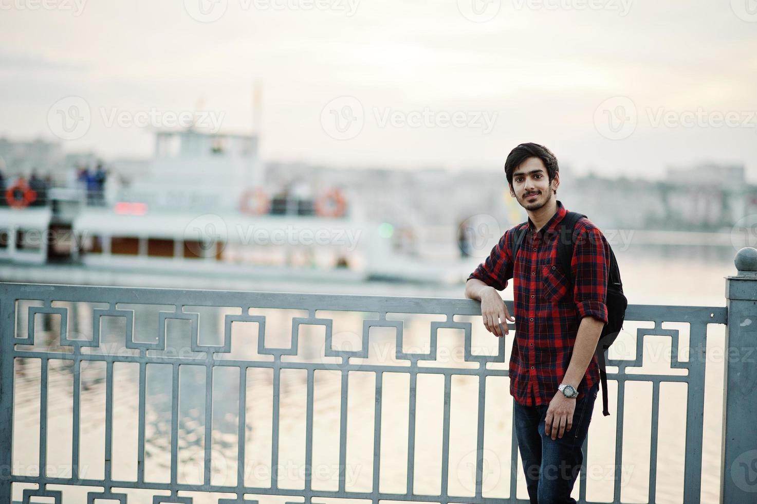 giovane studente indiano in camicia a scacchi e jeans con zaino in posa sulla città di sera contro il lago e la barca. foto