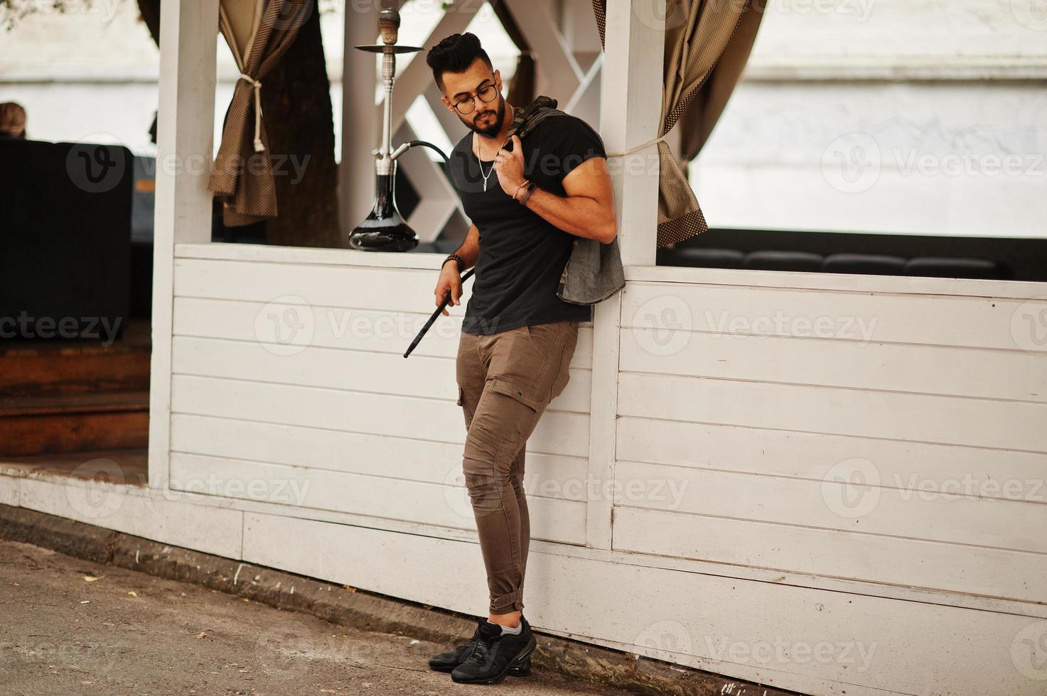 elegante barba uomo arabo in occhiali e t-shirt nera che fuma narghilè all'aperto. modello arabo che riposa. foto