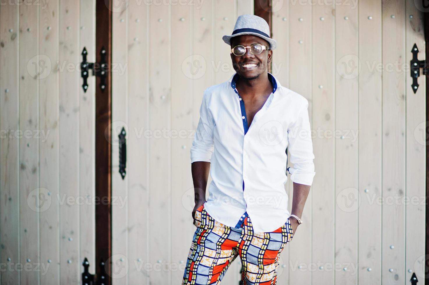 elegante uomo afroamericano in camicia bianca e pantaloni colorati con cappello e occhiali in posa all'aperto. ragazzo nero modello alla moda. foto