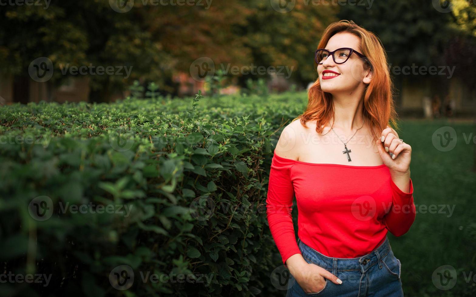 attraente donna dai capelli rossi in occhiali, indossare su camicetta rossa e gonna di jeans in posa al parco verde. foto