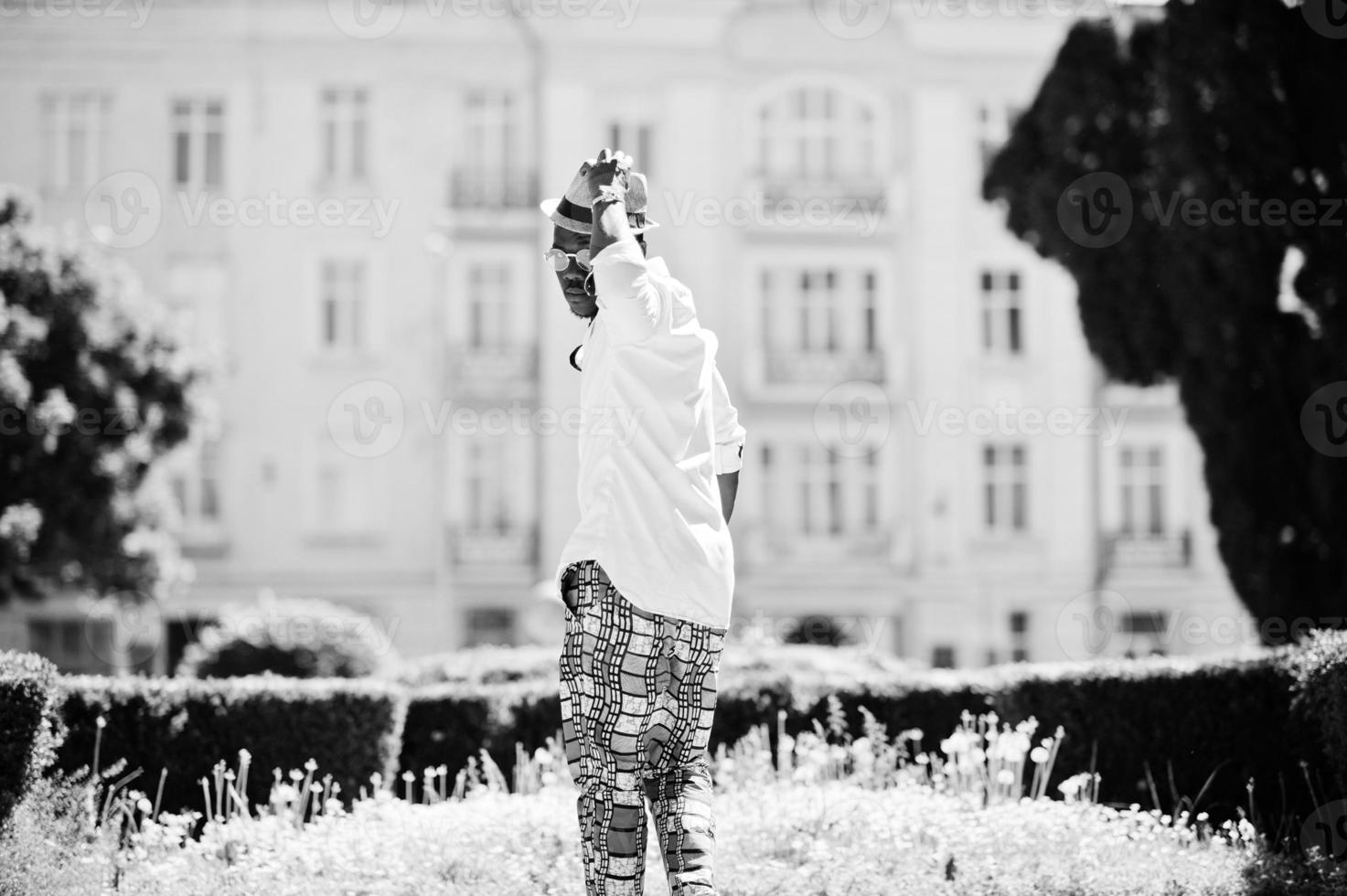 elegante uomo afroamericano in camicia bianca e pantaloni colorati con cappello e occhiali in posa all'aperto. ragazzo nero modello alla moda. foto