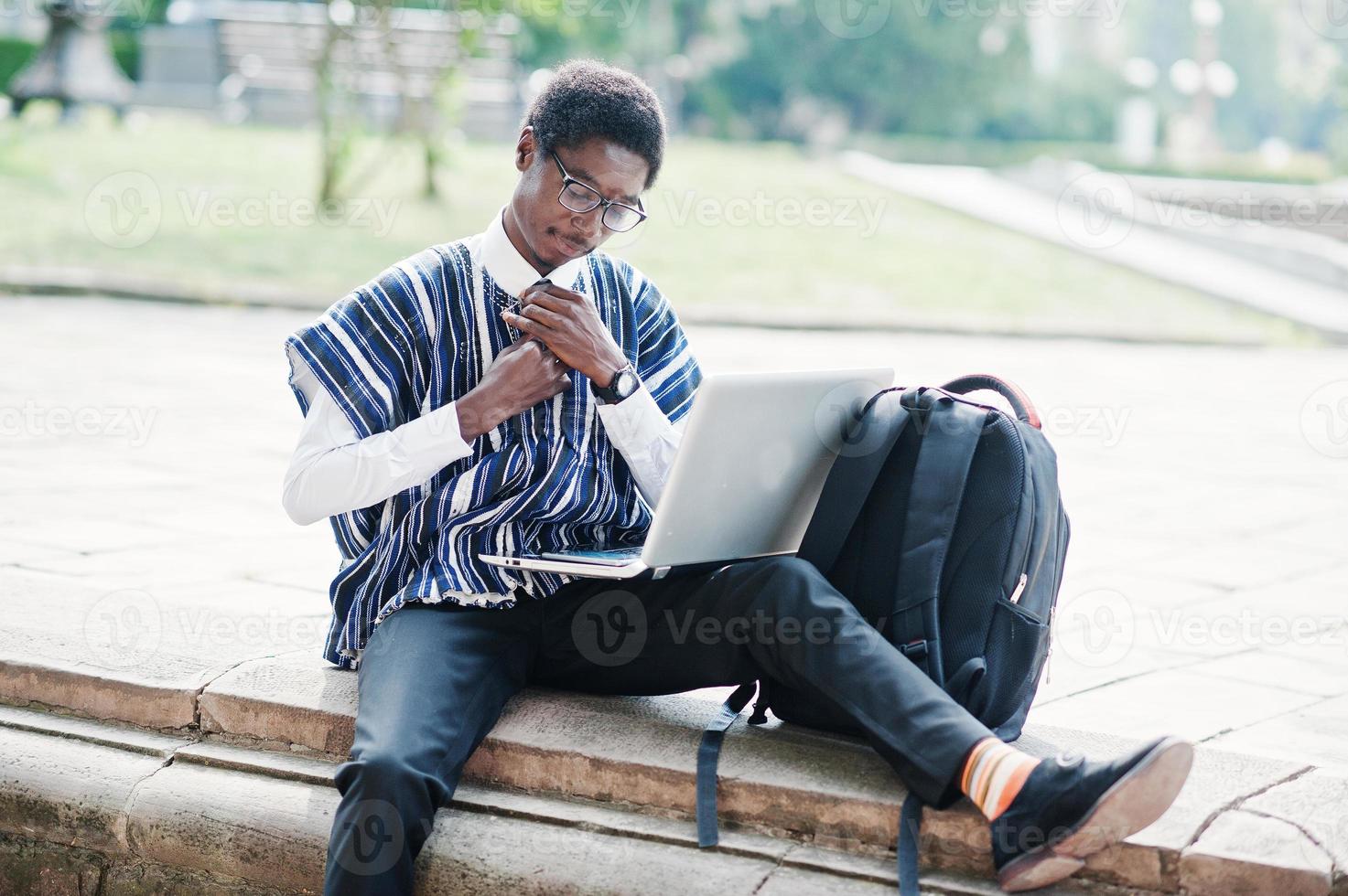 studente africano in abiti tradizionali e occhiali con zaino, lavorando al computer portatile all'aperto. foto