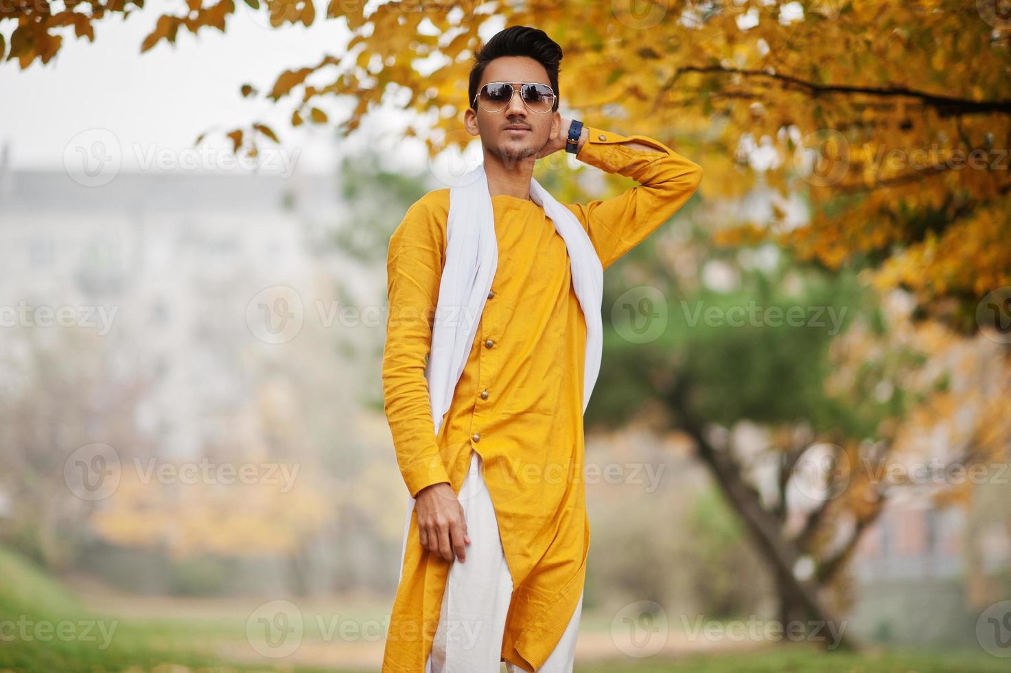 uomo indiano alla moda in abiti tradizionali gialli con sciarpa bianca, occhiali da sole in posa all'aperto contro l'albero delle foglie d'autunno. foto