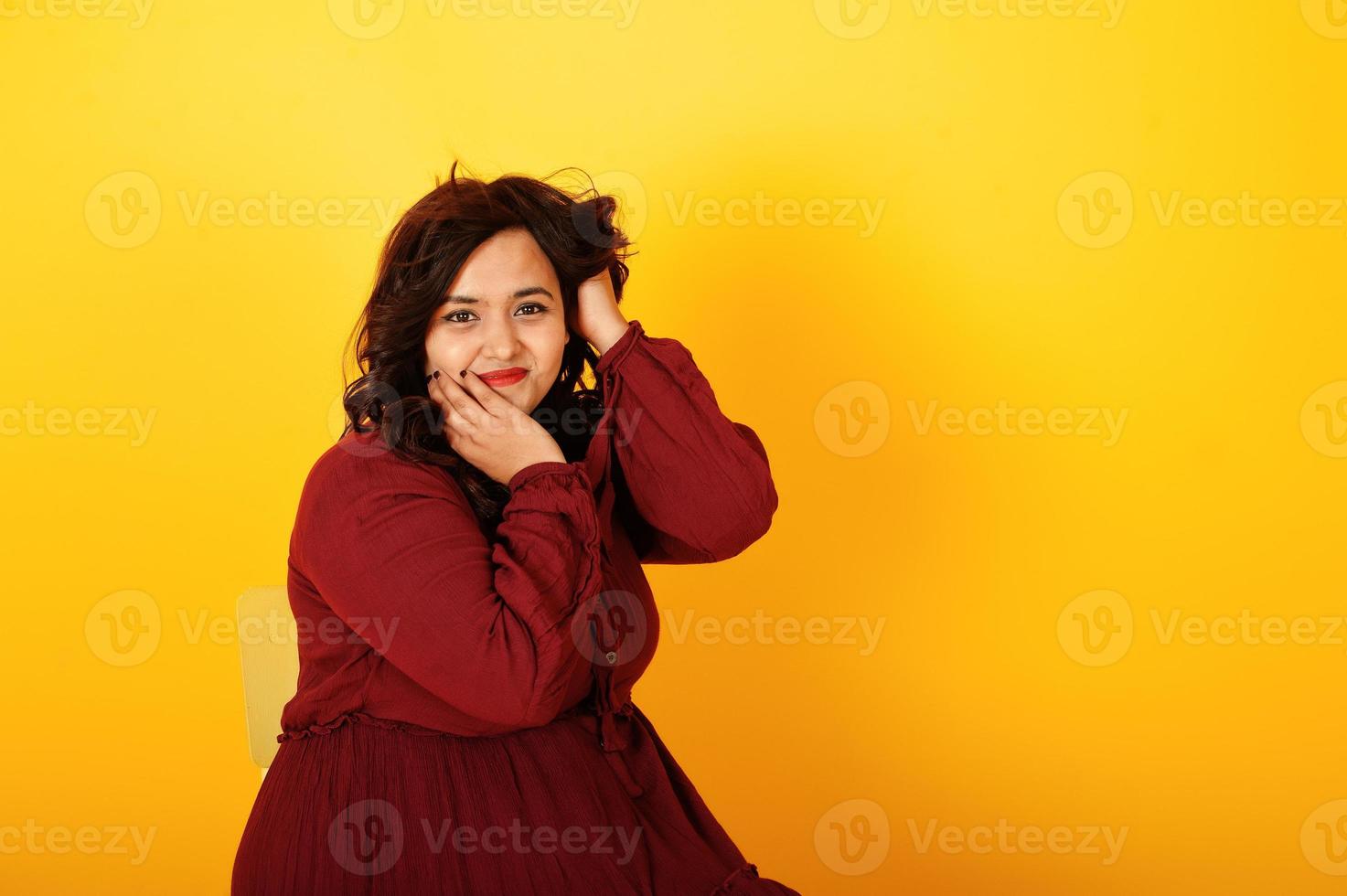 attraente donna del sud asiatico in abito rosso scuro posata in studio su sfondo giallo e seduta su una sedia. foto