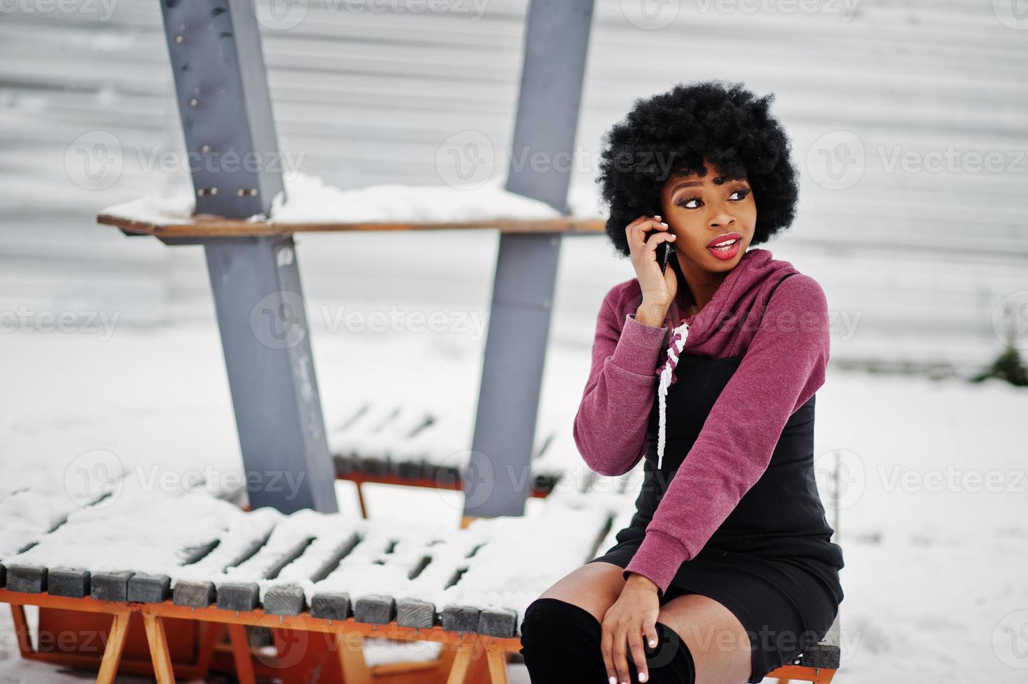 capelli ricci donna afroamericana in posa al giorno d'inverno, seduta su una panchina e parlando al cellulare. foto
