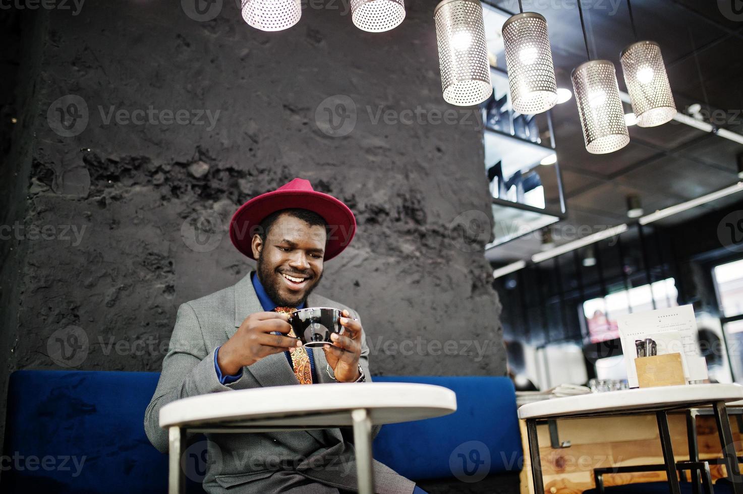 elegante uomo afroamericano modello in giacca grigia cravatta e cappello rosso bere caffè al bar. foto