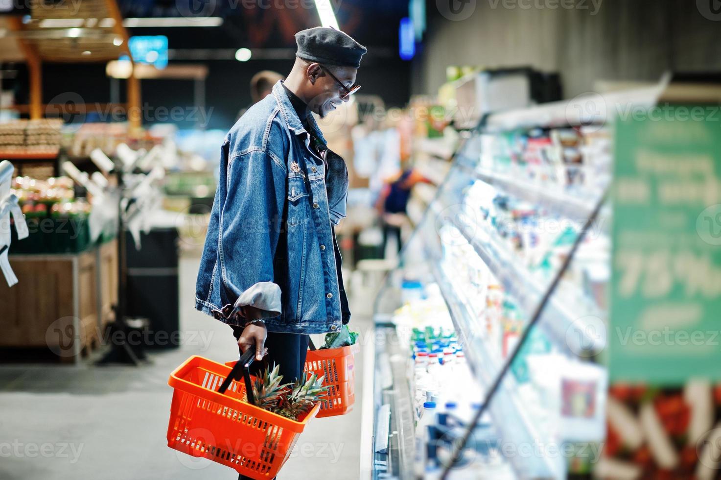 elegante uomo afroamericano casual con giacca di jeans e berretto nero con due cesti, in piedi vicino al frigorifero e shopping al supermercato. foto