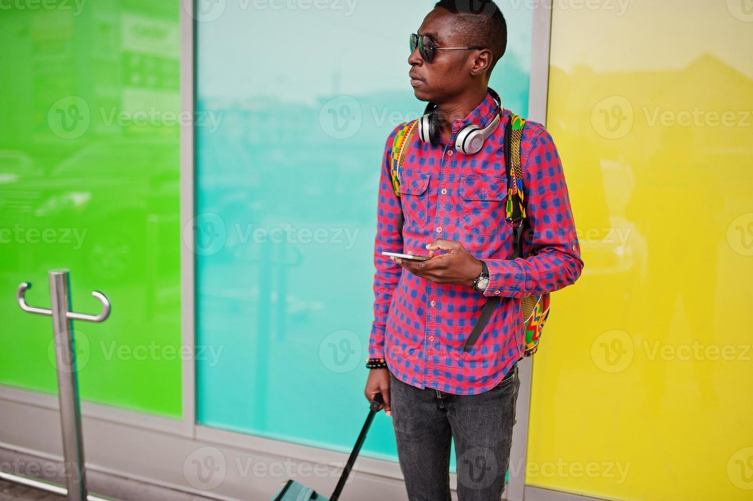 uomo afroamericano in camicia a scacchi, occhiali da sole e jeans con valigia e zaino. viaggiatore nero. foto