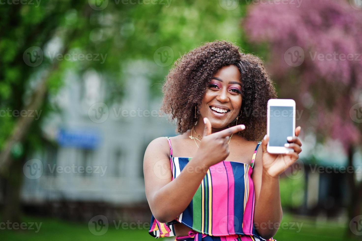 donna afroamericana alla moda in tuta a righe rosa posata in strada in fiore primaverile e mostra il dito sullo schermo del telefono cellulare. foto