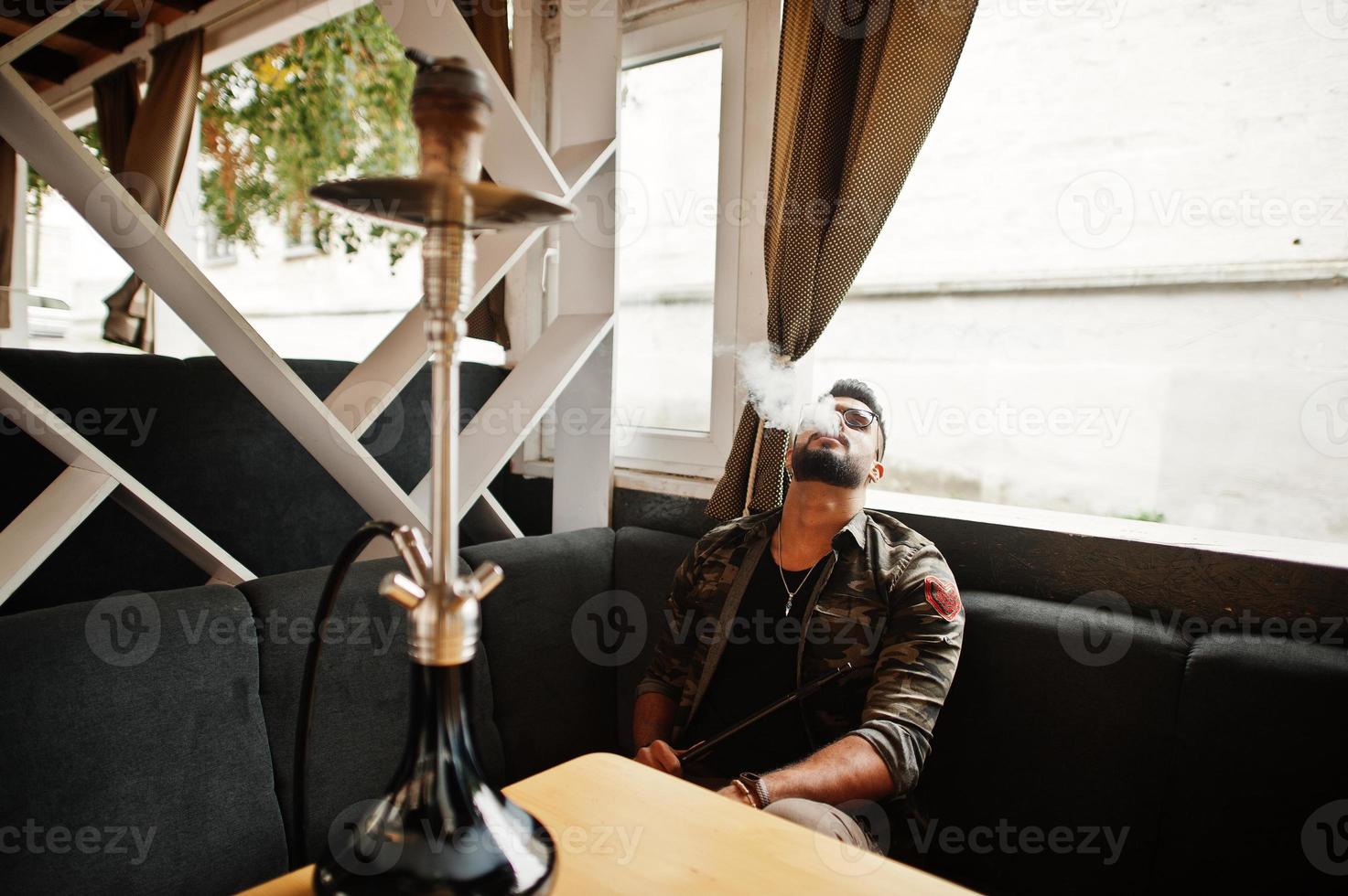 elegante barba uomo arabo con gli occhiali e giacca militare che fuma narghilè al bar di strada. modello arabo che riposa. foto