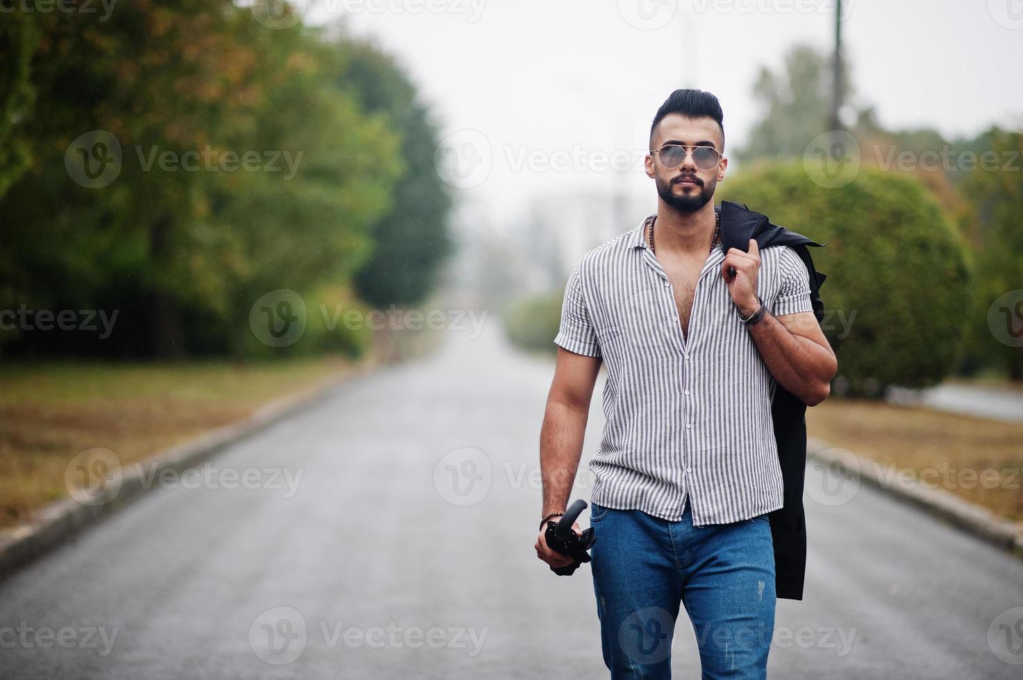 l'uomo alla moda con barba araba alta indossa camicia, jeans e occhiali da sole che camminano al parco con ombrello e cappotto a portata di mano. foto