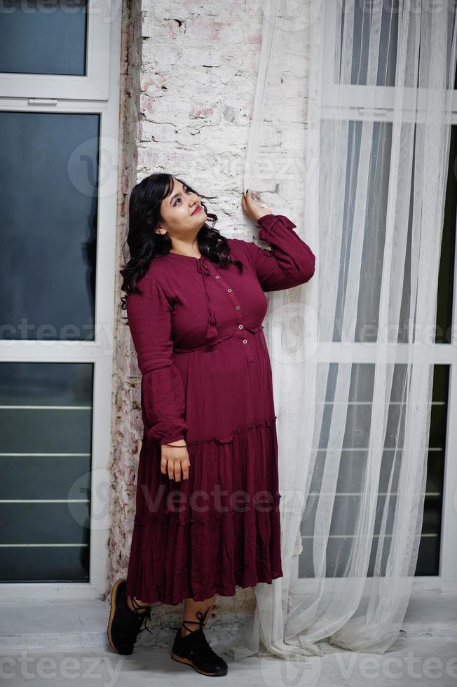 attraente donna del sud asiatico in abito rosso intenso poste in studio contro le finestre nella notte. foto