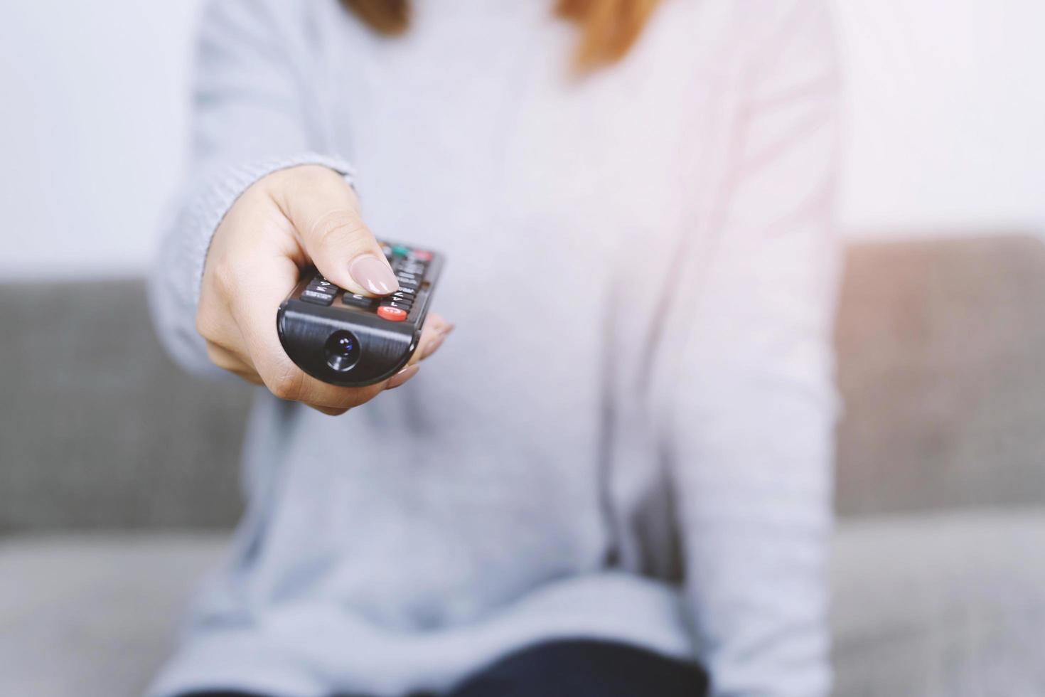 primo piano telecomando della televisione nelle mani casual della camicia gialla femminile giovane che puntano al televisore e lo accendono o lo spengono. seleziona il canale guardando la tv sul divano di casa in soggiorno rilassati. foto