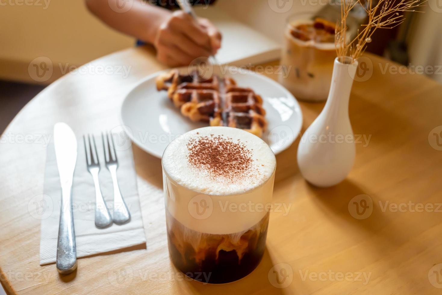 un pasto mattutino della domenica al bar, due tazze di caffè, latte macchiato al miele e eispanner americano serviti con waffle su piatto, decorato con vaso. foto