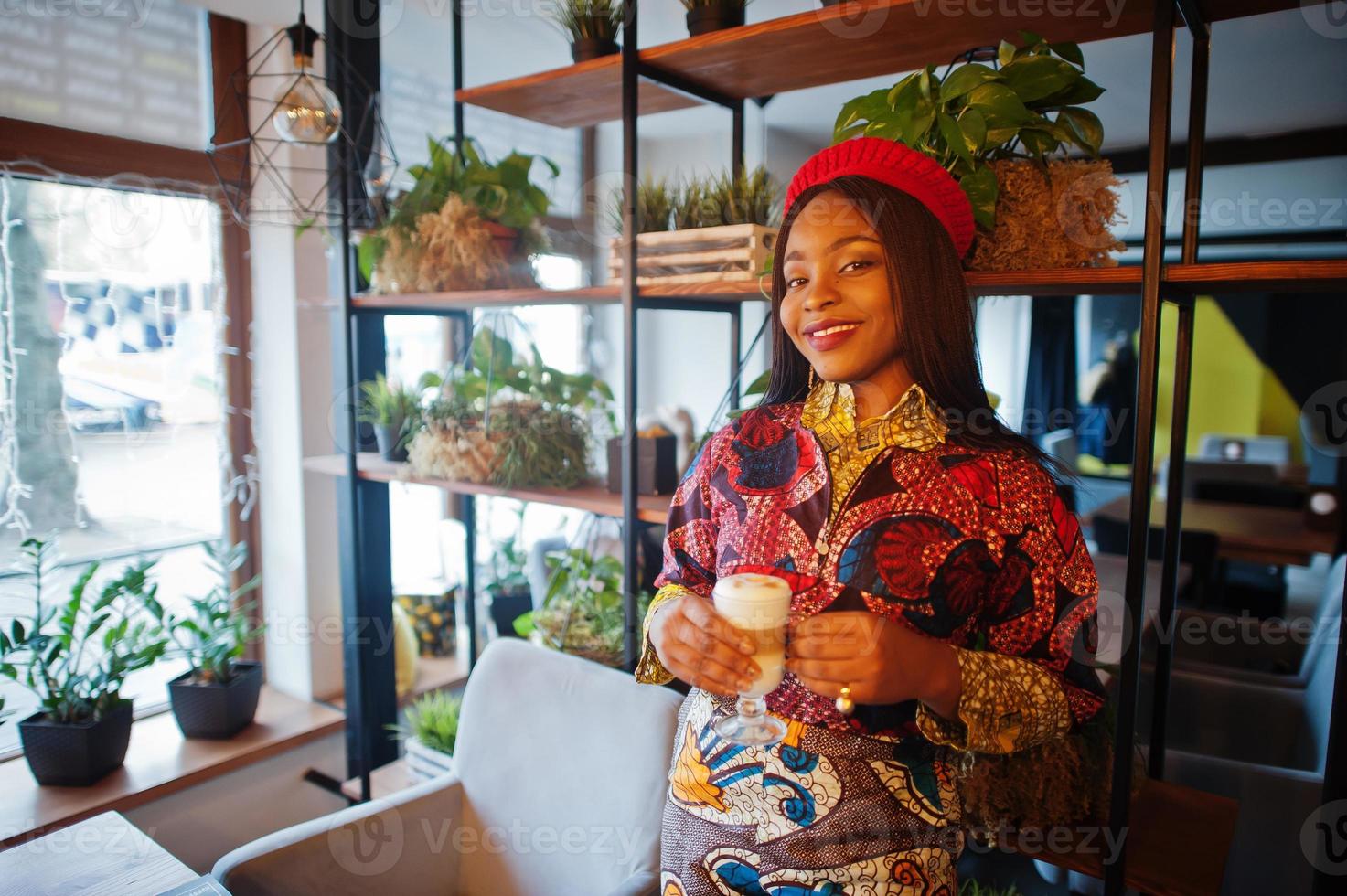 entusiasta donna afroamericana in abito colorato alla moda con berretto rosso che si rilassa in un accogliente caffè con una tazza di cappuccino a portata di mano. foto