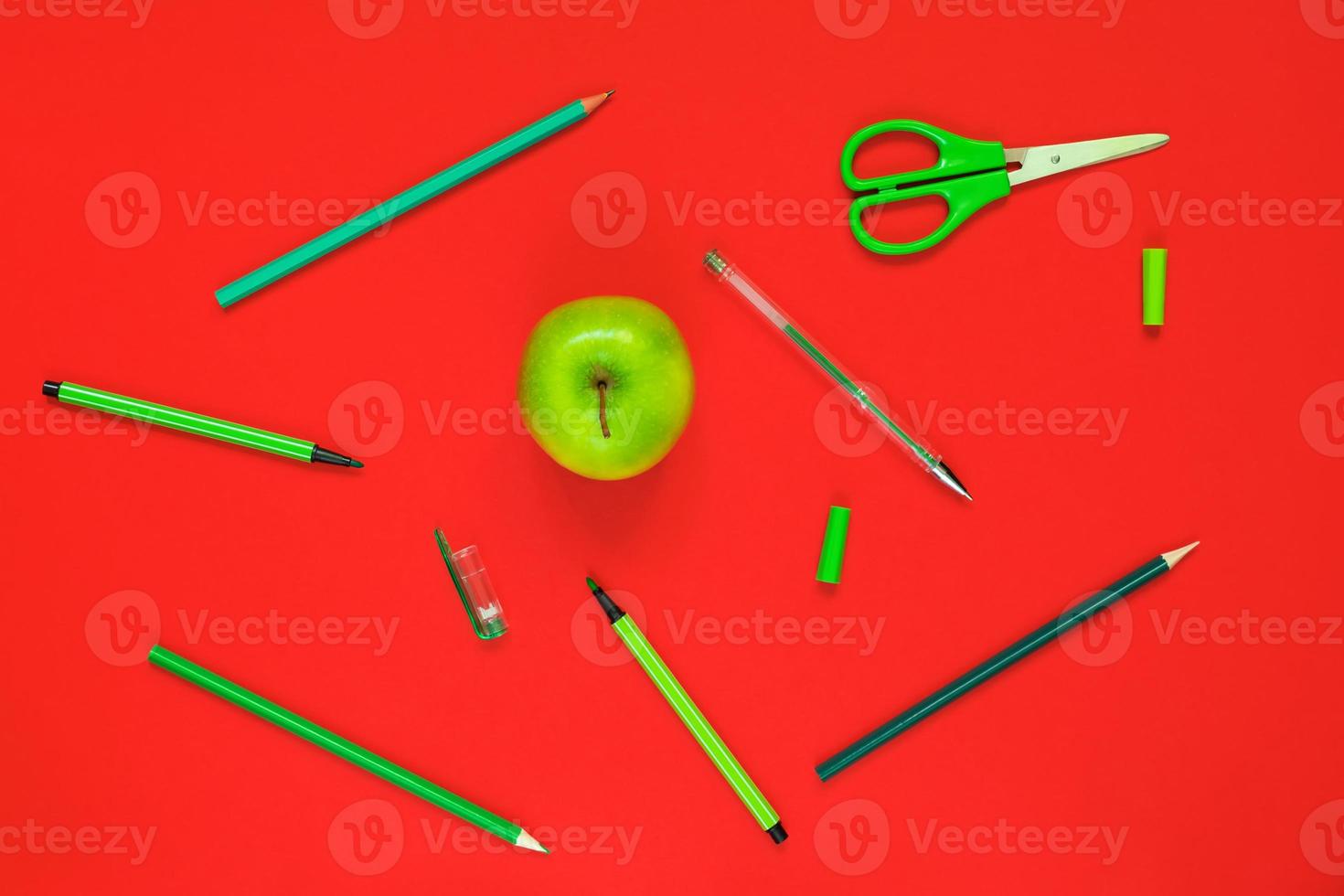 disposizione piatta creativa di materiale scolastico - penne verdi, matite, forbici e una mela su sfondo rosso foto