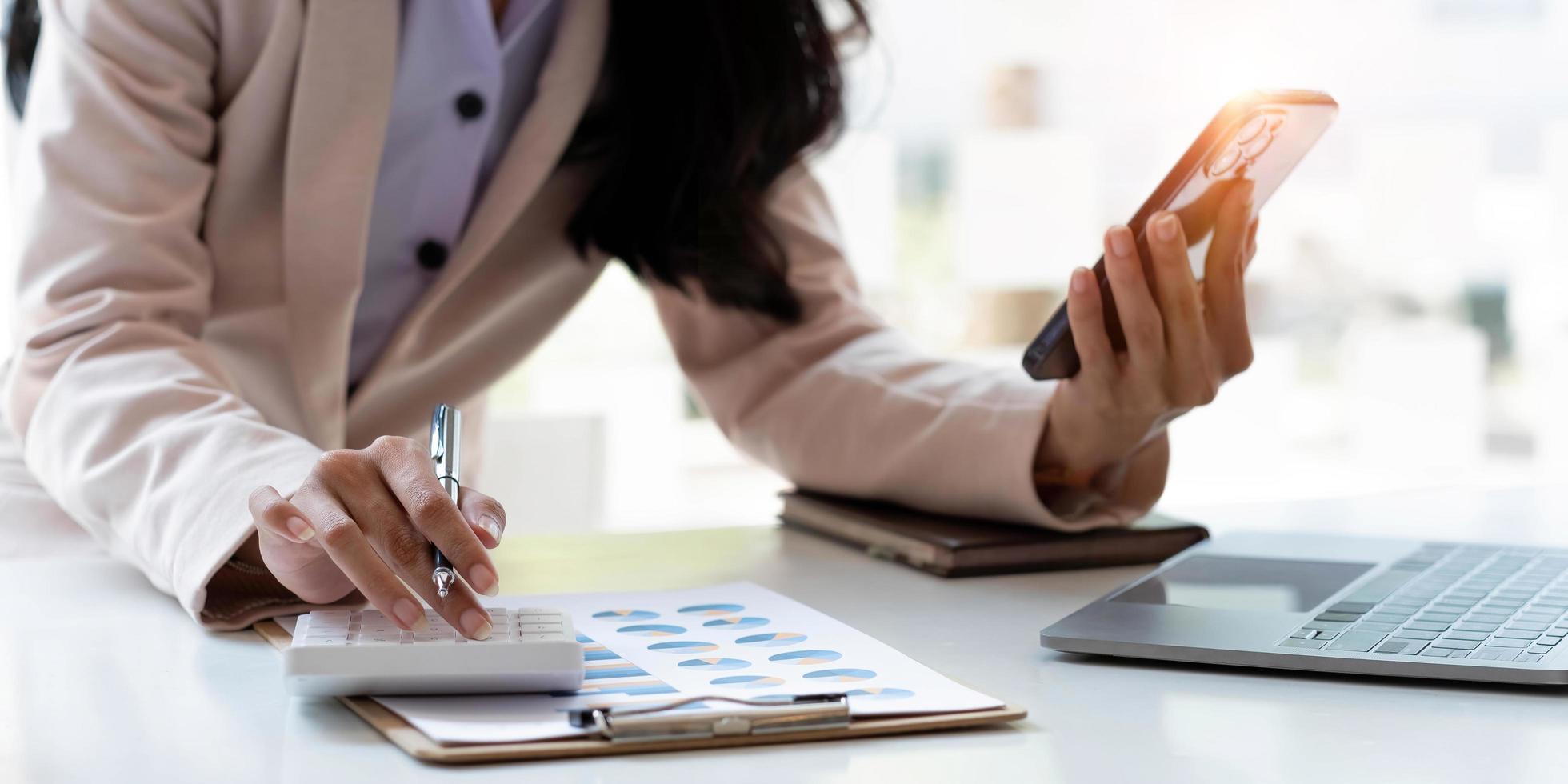 attività commerciale donna o contabile mano utilizzando calcolatrice, Lavorando su mobile inteligente Telefono e il computer portatile computer con documentario dati grafico carta su il tavolo a moderno casa ufficio. in linea marketing concetto. foto