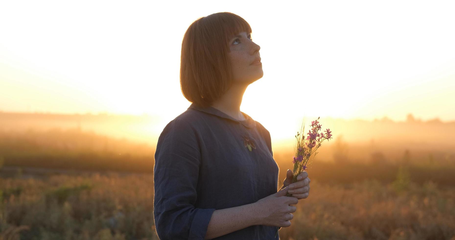 giovane testa Rossa donna nel bellissimo boho vestito rilassante nel il campo durante nebbioso tramonto, femmina all'aperto con mazzo nel mani foto