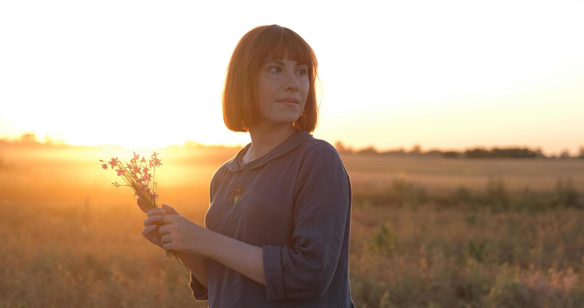 giovane testa Rossa donna nel bellissimo boho vestito rilassante nel il campo durante nebbioso tramonto, femmina all'aperto con mazzo nel mani foto