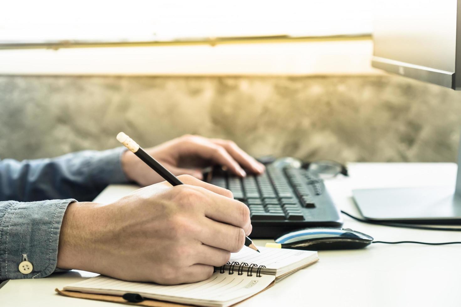 vicino su di maschio mani scrittura alcuni dati nel taccuino e utilizzando computer su il scrivania foto