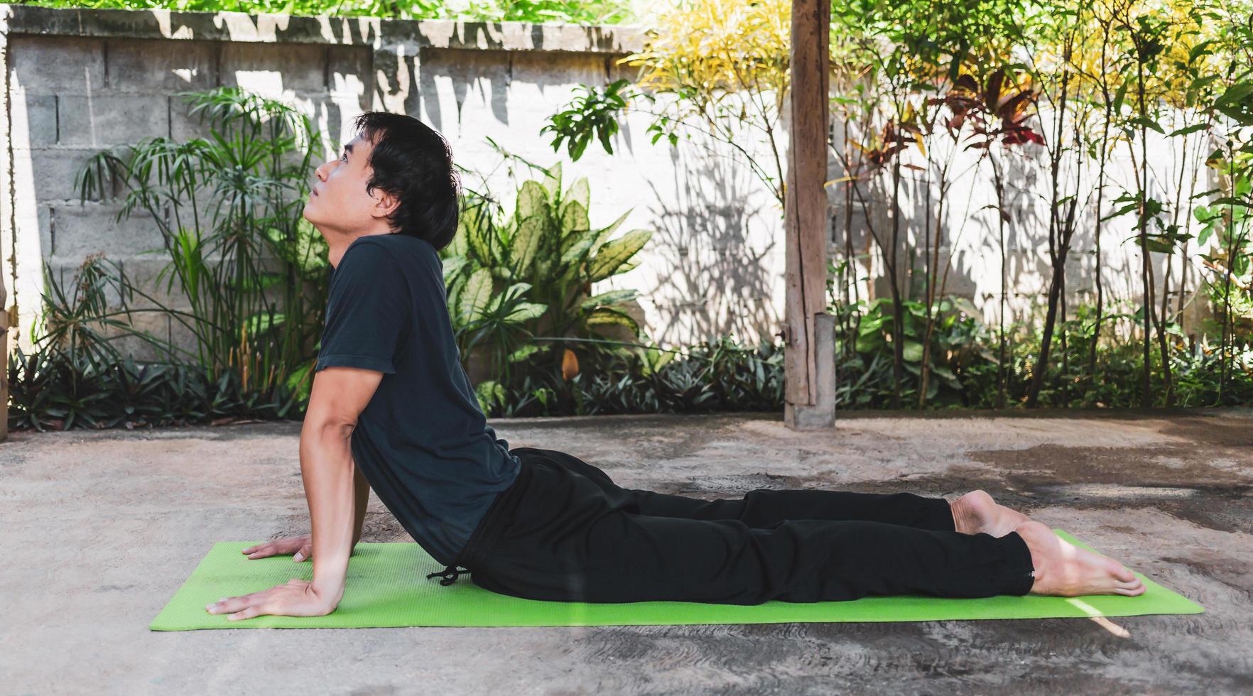 asiatico uomo nel casuale attrezzatura praticante yoga nel cobra posa su il verde yoga stuoia nel il giardino a casa. salutare stile di vita e salutare vivente concetto. foto