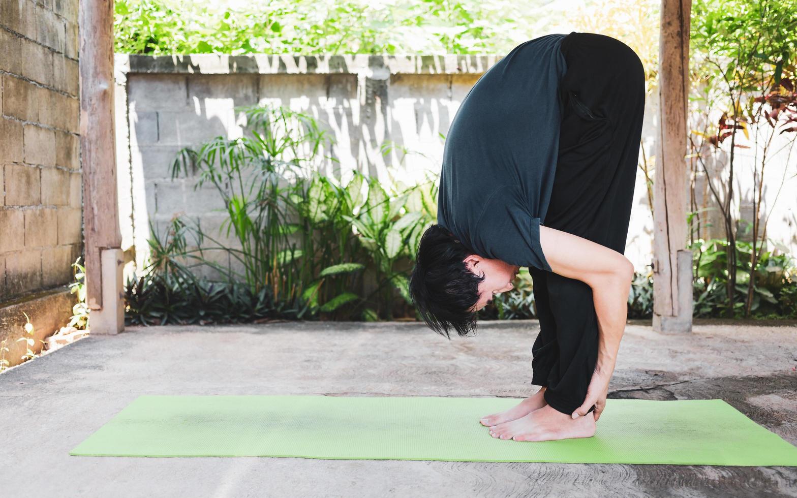 salutare vivente concetto di giovane asiatico uomo praticante yoga asana Uttanasana - in piedi inoltrare piegare posa, Lavorando fuori, pose su un' verde yoga stuoia. all'aperto esercizio nel il giardino. salutare stile di vita foto