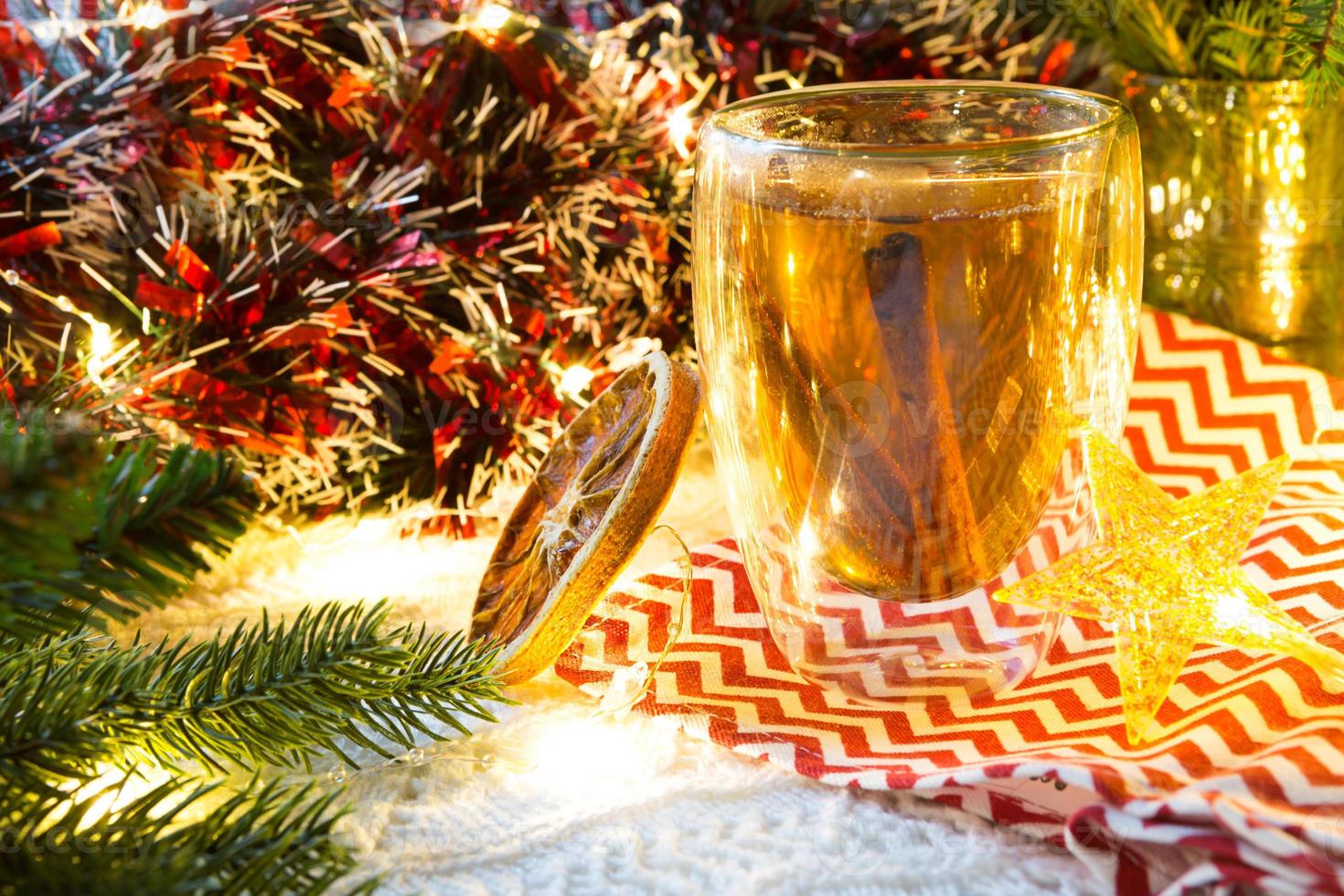 trasparente a doppia parete bicchiere bicchiere con caldo tè e cannella bastoni su il tavolo con Natale arredamento. nuovo anni atmosfera, fetta di secco arancia, ghirlanda e orpelli, abete rosso ramo, accogliente foto