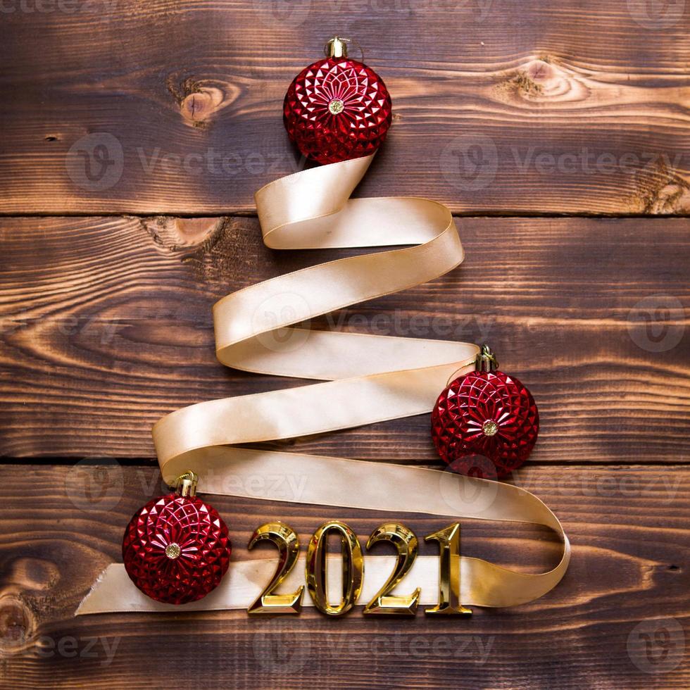 un albero di natale fatto di nastro dorato con i numeri 2021 è decorato con palloncini rossi su fondo di legno scuro. disteso. spazio per il testo. capodanno, cappello da Babbo Natale. foto