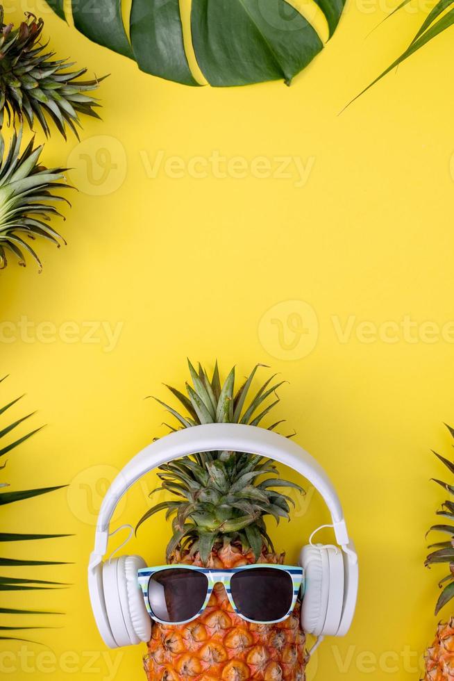 divertente ananas che indossa una cuffia bianca, ascolta musica, isolata su sfondo giallo con foglie di palma tropicali, vista dall'alto, concetto di design piatto. foto