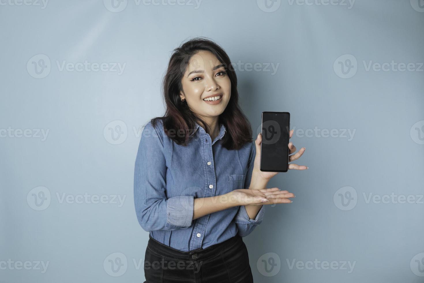 eccitato asiatico donna indossare blu camicia puntamento a il copia spazio su sua smartphone, isolato di blu sfondo foto