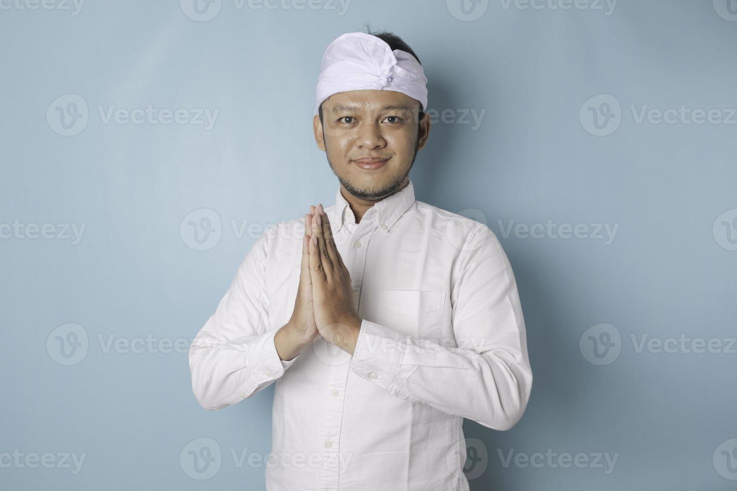 sorridente giovane balinese uomo indossare udeng o tradizionale fascia per capelli e bianca camicia Esprimere a gesti saluto o namaste isolato al di sopra di blu sfondo foto