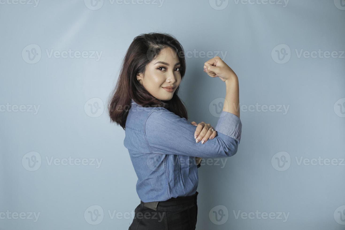 eccitato asiatico donna indossare un' blu camicia mostrando forte gesto di sollevamento sua braccia e muscoli sorridente con orgoglio foto