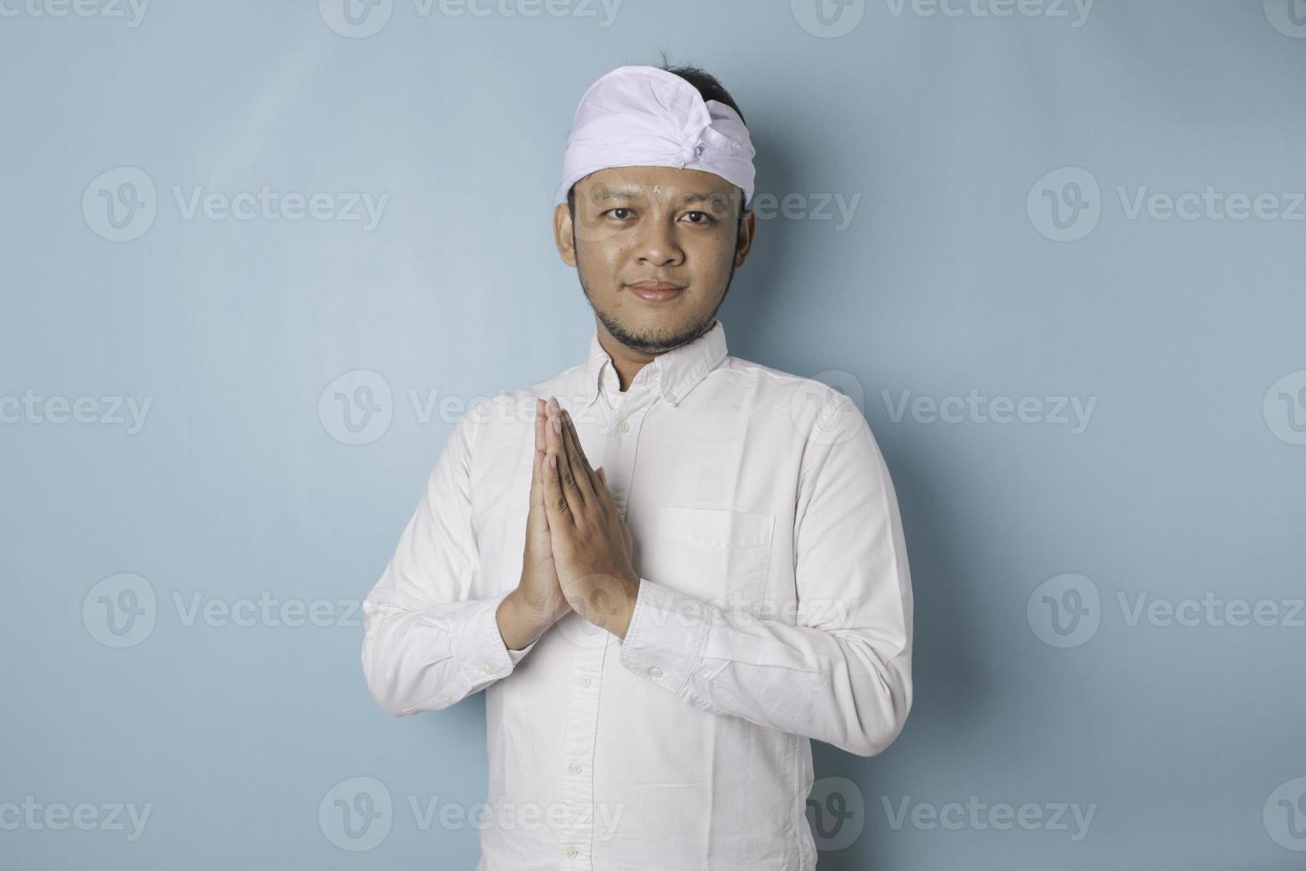 sorridente giovane balinese uomo indossare udeng o tradizionale fascia per capelli e bianca camicia Esprimere a gesti saluto o namaste isolato al di sopra di blu sfondo foto