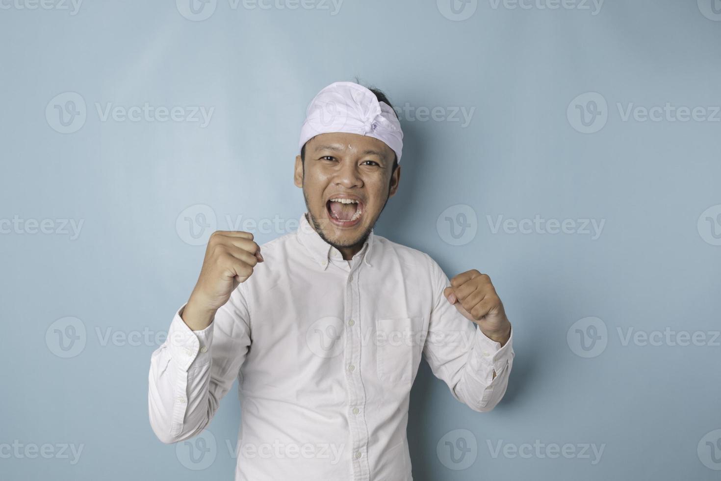 un' giovane balinese uomo con un' contento riuscito espressione indossare udeng o tradizionale fascia per capelli e bianca camicia isolato di blu sfondo foto