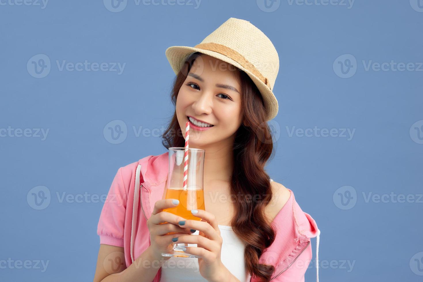 ritratto di contento giovane asiatico donna indossare cannuccia cappello e potabile frutta succo al di sopra di blu sfondo. foto