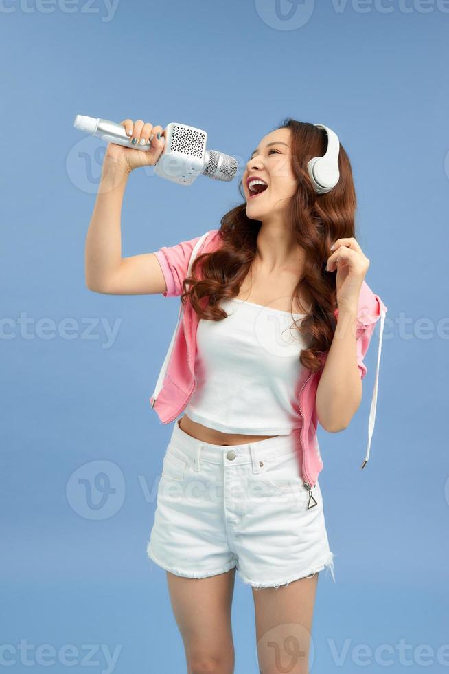 contento giovane asiatico donna cantando con microfono cantare e cuffie al di sopra di blu sfondo. foto