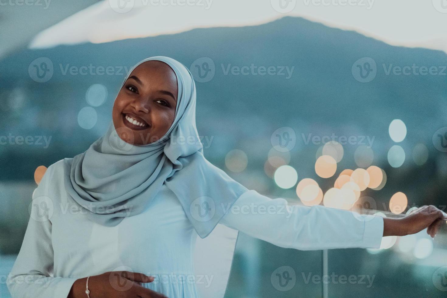 africano musulmano donna nel il notte su un' balcone sorridente a il telecamera con città bokeh luci nel il sfondo. foto