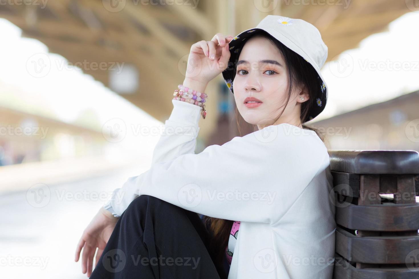 asiatico bellissimo ragazza nel bianca lungo manica camicia e cappello sedersi nel il treno stazione per aspettare treno arrivato. foto