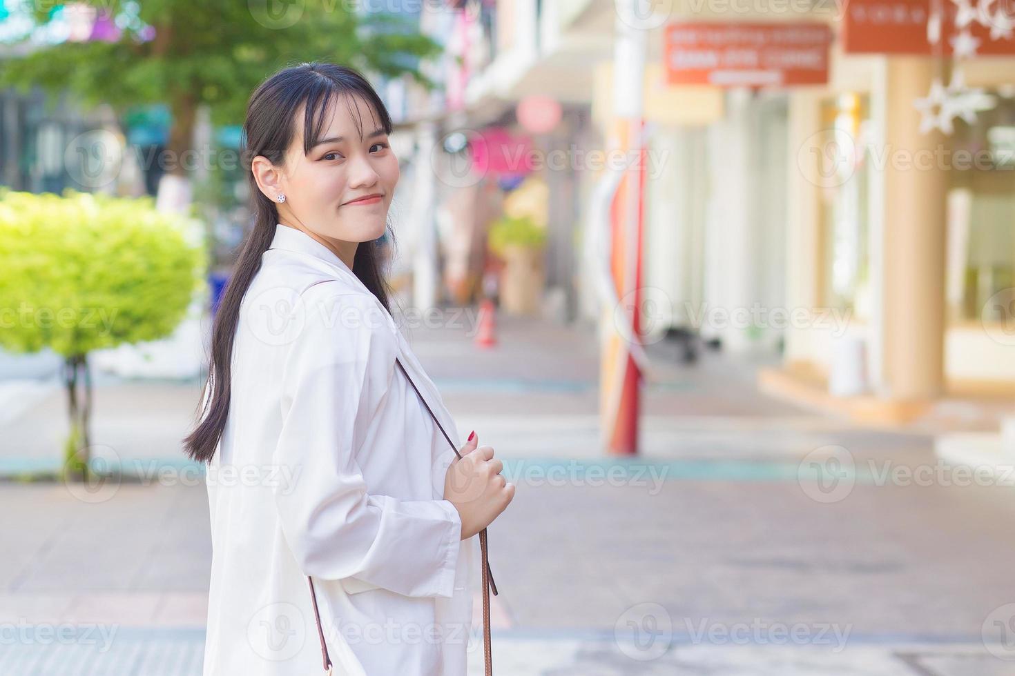 fiducioso giovane attività commerciale asiatico Lavorando femmina chi indossa un' bianca camicia e spalla Borsa sorrisi felicemente mentre lei è a piedi per opera a ufficio nel il città con un' negozio edificio nel il sfondo. foto