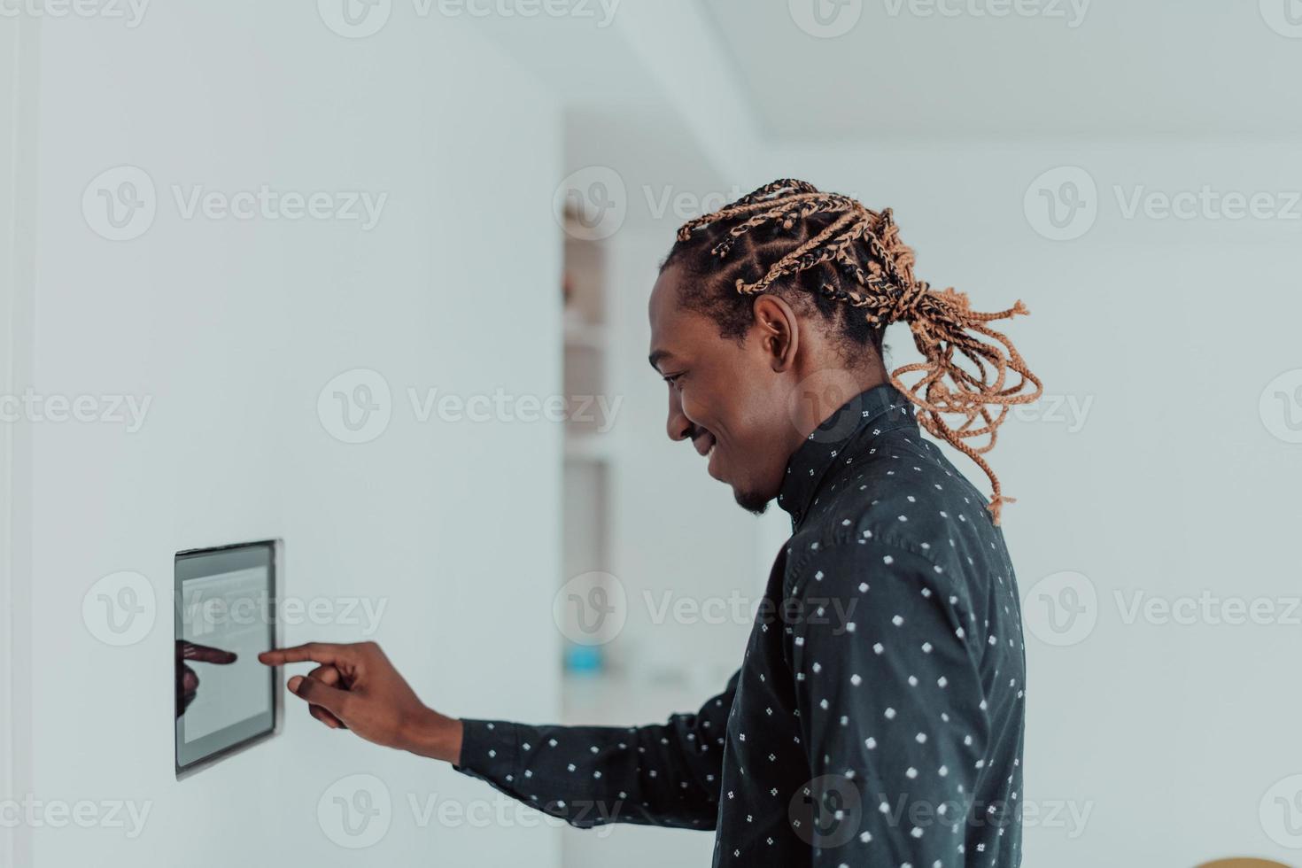 sorridente africano americano uomo utilizzando moderno inteligente casa sistema, controllore su parete, positivo giovane uomo commutazione temperatura su termostato o attivazione sicurezza allarme nel appartamento foto